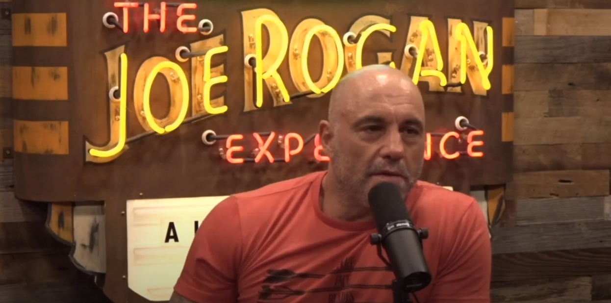 Joe Rogan in orange shirt, speaking into microphone