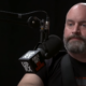 Tom Segura in front of a microphone against a dark background