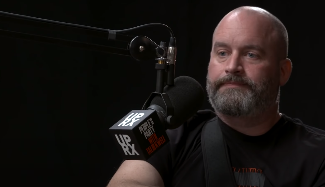 Tom Segura in front of a microphone against a dark background