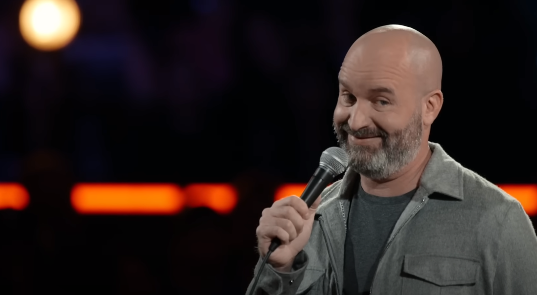 Tom Segura smiling with a slightly tilted head, holding a microphone