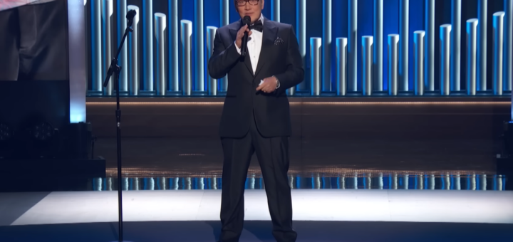 Rob Schneider in a black suit standing on the stage and holding the microphone