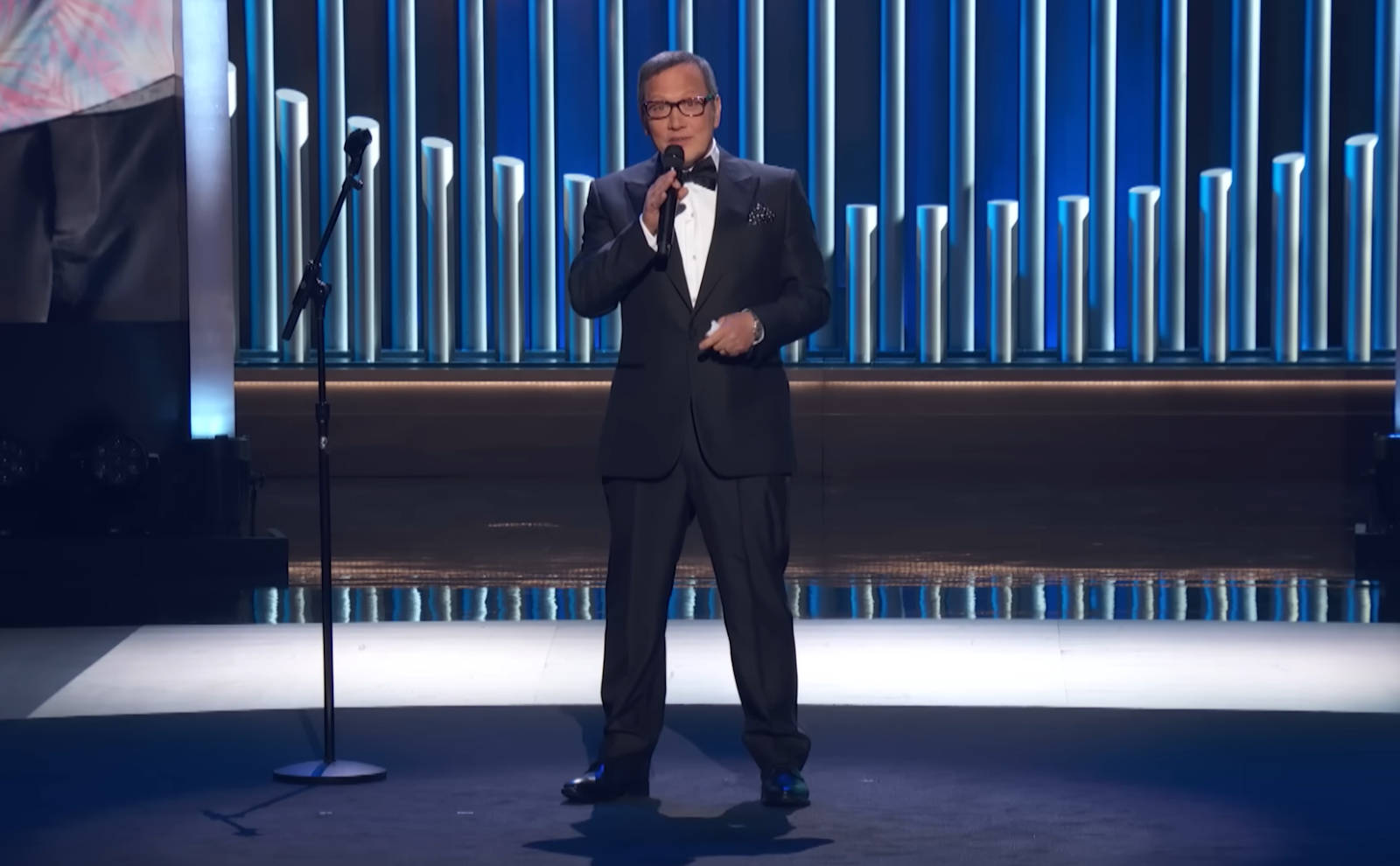 Rob Schneider in a black suit standing on the stage and holding the microphone