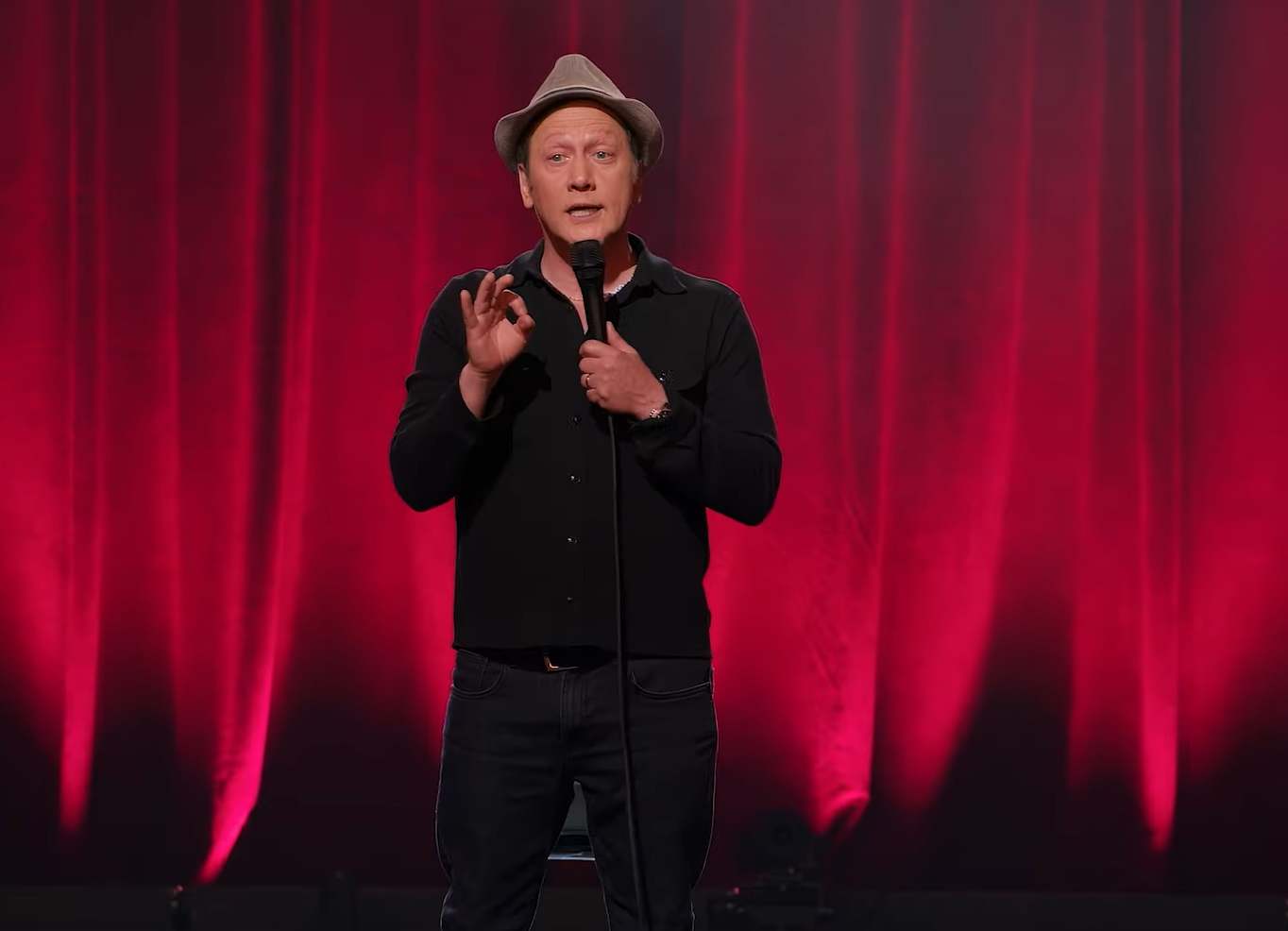 Rob Schneider in hat and black clothes standing on stage with red curtains behind