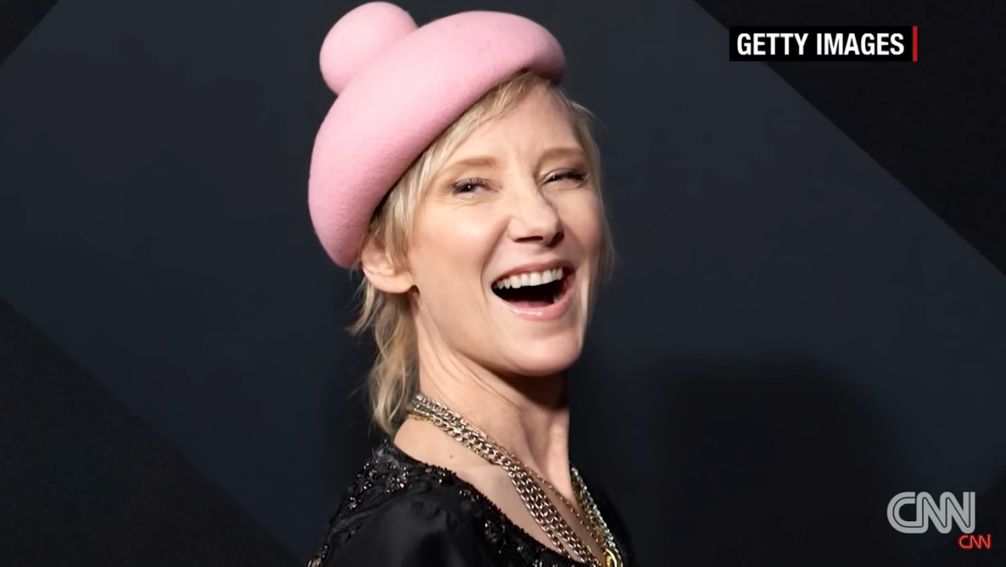 anne heche in pink hat standing and smiling near black wall