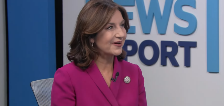 Joy Hofmeister in a dark pink suit sitting in a chair, smiling and looking at the side