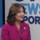 Joy Hofmeister in a dark pink suit sitting in a chair, smiling and looking at the side