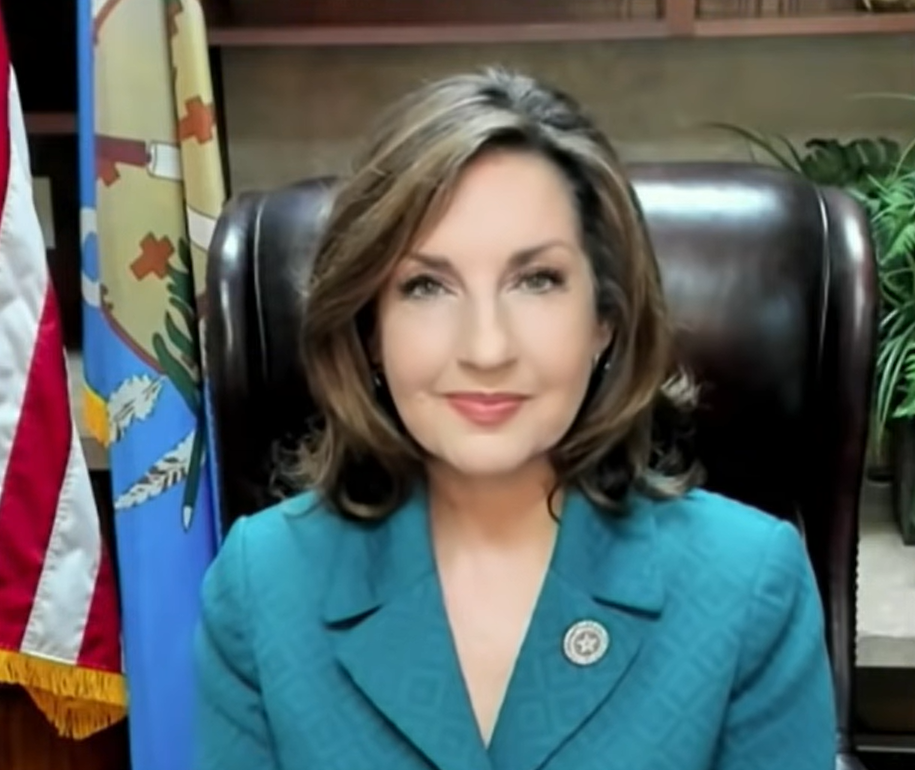 Joy Hofmeister in a blue jacket sitting in a leather brown chair, flags behind her