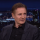 Liam Neeson in a suit against a cityscape night background